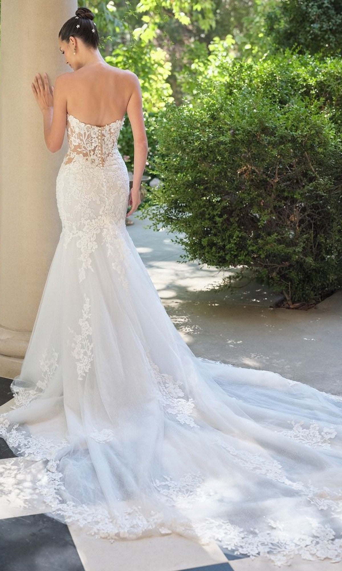 A woman stands outdoors in the Andrea and Leo WL065 long wedding dress, a strapless lace mermaid gown with a long train, facing away as she rests one hand on a column.